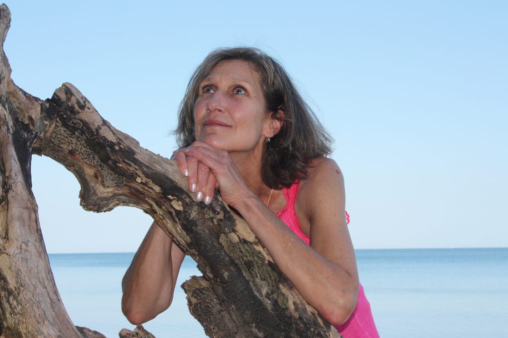 Andrea Geise am Strand, nachdenklich an einen Baumstamm gelehnt.